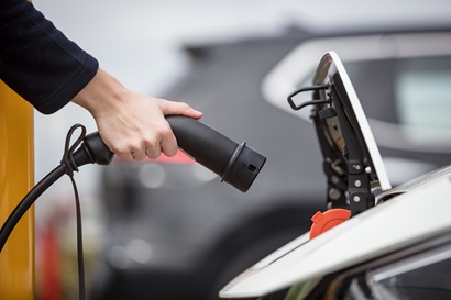 Photograph of an arm and hand holding an electric vehicle charging plug next to an EV.