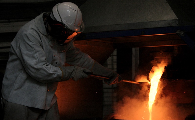Heavily protected helmeted worker beside furnace with molten metal flowing out of chamber
