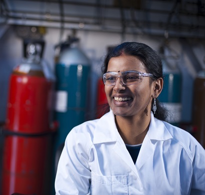 Female researcher wearing safety glasses and white lab coat, smiling and looking away from camera, with a row of gas bottles behind her, in the gas to liquids laboratory.