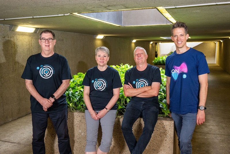 4 people (3 men and 1 woman) wearing dark coloured T-shirts are in an undercover area. The man and woman in the middle are sitting on the edge of a raised garden bed and the two men on the outside are standing. All are smiling at the camera.