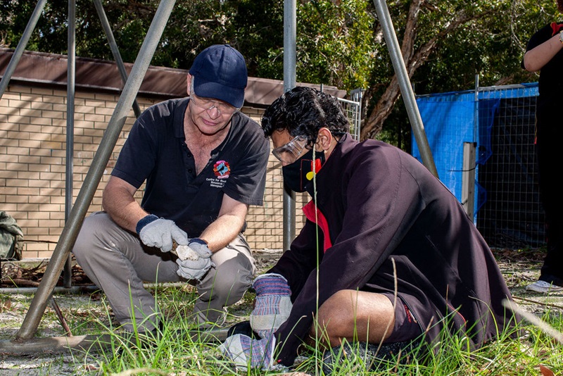Teacher and Indigenous student. Man in a navy cap, gloves and safety goggles is doing a demonstration for a student who is seated next to him wearing gloves, safety goggles and a mask with the Australian Aboriginal flag on it.
