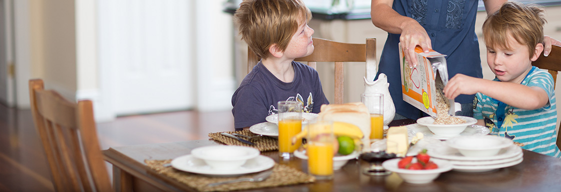 Woman pouring Goodness Superfoods brand cereal for her two children at the breakfast table