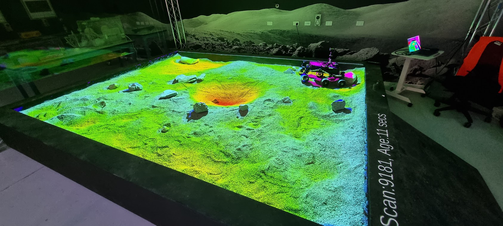 A picture of a coloured digital projection on top of a large sand pit with rocks and a small rover in the far right corner.