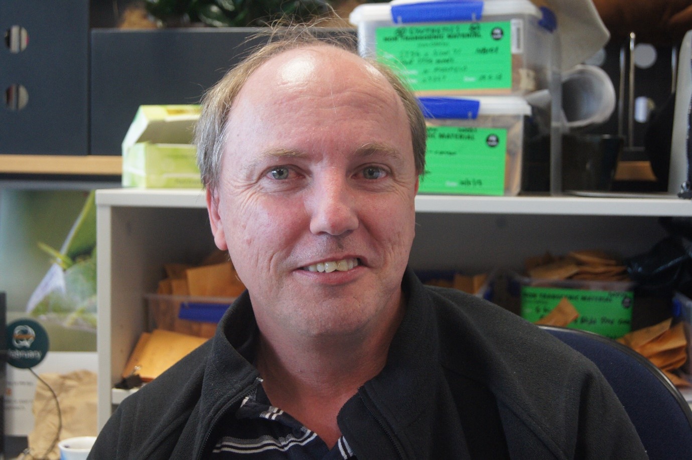 A man smiles for the camera in front of a shelf crammed with labelled plastic containers.