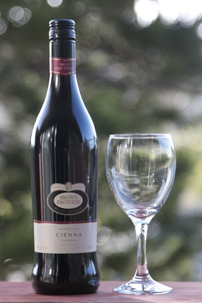 Close shot of a bottle of Brown Brothers Cienna wine beside an empty wine glass, with green folliage background