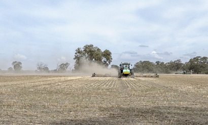 Farm machinery ploughs through an open field of stubble, throwing up dust behind it