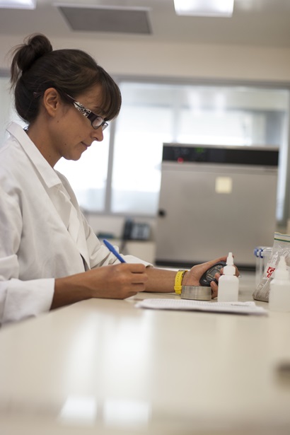 A female CSIRO scientist in a white lab coat and glasses sitting at a white lab bench holding a blue pen and black stopwatch watching a small tin of soil on the bench