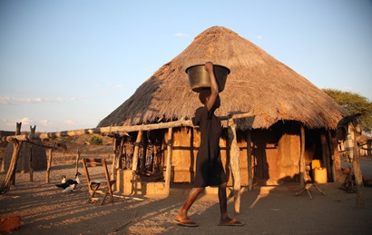 Woman walks past a simple hut in a village in Africa carrying a container on her head