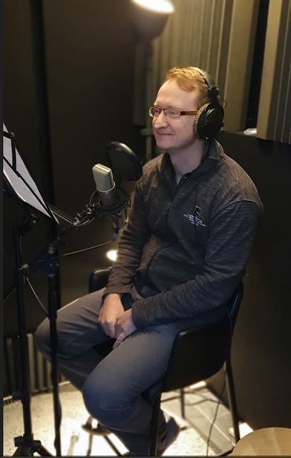 Jon sits in a recording studio. He is wearing headphones and facing a mike. 