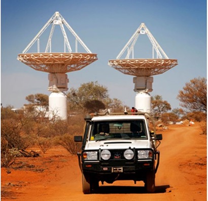 The accompanying image contains a photo of a large 4WD in the foreground and it is driving on a dirt road on Wajarri land in Western Australia. In the background are medium sized shrubs and one of our large ASKAP satellites.