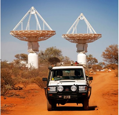 The accompanying image contains a photo of a large 4WD in the foreground and it is driving on a dirt road on Wajarri land in Western Australia. In the background are medium sized shrubs and one of our large ASKAP satellites.