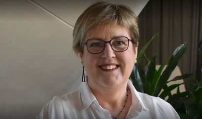 This image is a photo of Deb Miller from the shoulder height up. She is wearing a white collared shirt, deep red lipstick, glasses, earrings and has blonde short hair. She is smiling. The image background is a tiled wall with a green plant to the right.