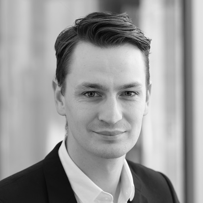 This is an portrait photo of Dominic Banfield. The photo is of his shoulders up and he is facing the camera from an angle. He is wearing an black suit with white collared shirt. He has short hair.