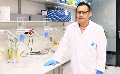 This image is a photo of Warish Ahmed standing in a CSIRO laboratory. He is wearing a lab coat, blue gloves and glasses.