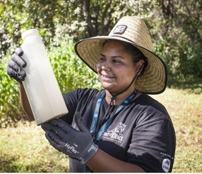 This is a photo of Kaylytah on Country. She is wearing a CSIRO shirt, large straw hat, gloves, a CSIRO lanyard. In the background there is grass and shrubs.  