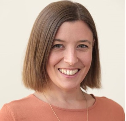 This image is a portrait photo of Lauren from the shoulders up. She has shoulder length hair which she is wearing out. She is smiling and is wearing a peach coloured shirt. She is standing in front of a cream wall.