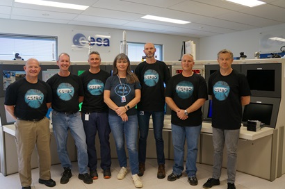 This image is a photo of the New Norcia Operations Team. There are 7 people in the photo and they are standing in the New Norcia Ground Station central workspace in front of their desks and dashboards. They are wearing matching CSIRO shirts and are all smiling.