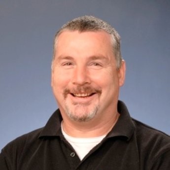 This image is a portrait photo of David. He is wearing a black polo shirt with a white top underneath. The background is blue and it is a professional photo that has been taken.