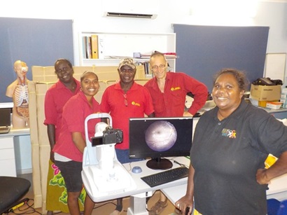 This image is a photo of the Remote Eye-care Research and Delivery Team in an office setting. There are 5 people in the photo and they are standing in front of a desk which has their desktop retinal camera on it. The people are all smiling.
