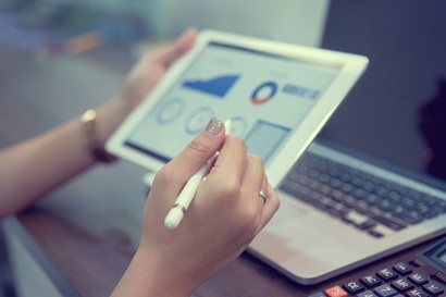 The accompanying image is a close up photo of a tablet screen with a person holding a pen. The screen shows tables and data visually presented but this is out of focus. 