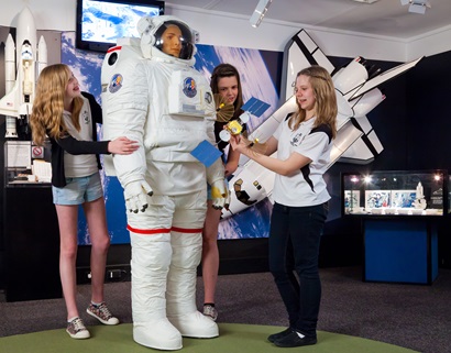 Three female school students looking at a mannequin in a NASA space suit in the foreground, models of space shuttles in the background