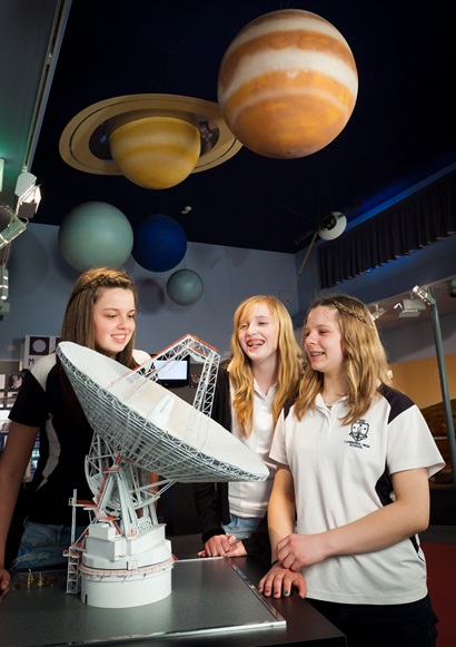 Three female students looking at a white scale model of an antenna, models of planets hang from the ceiling above.