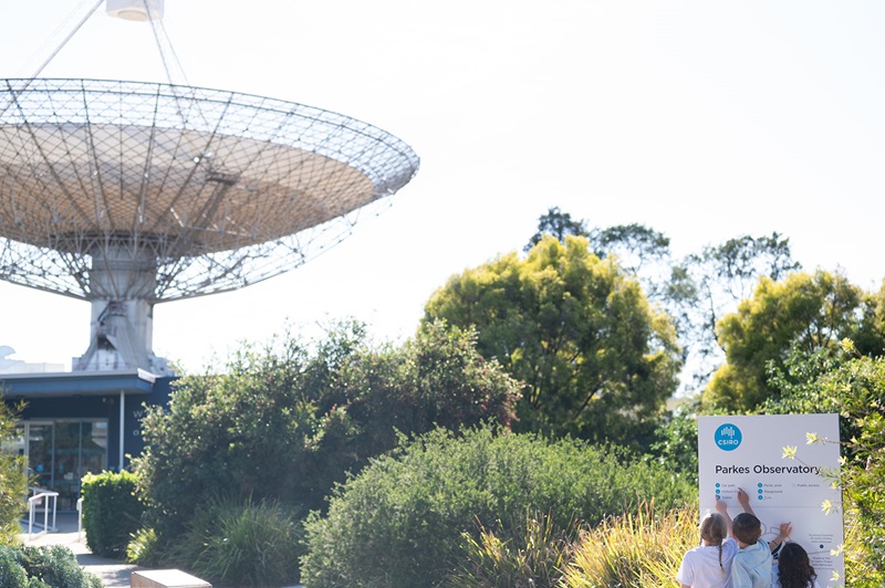 Children point to a sign that says Parkes Observatory. A radio telescope can be seen in the background.