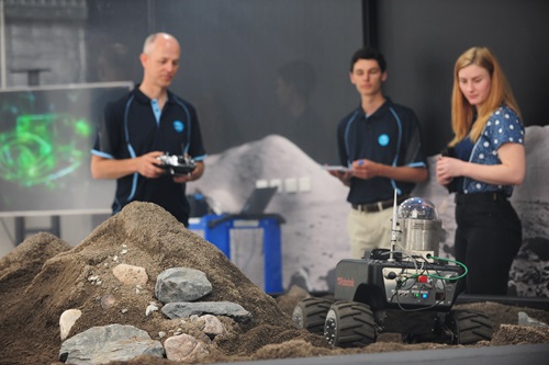 One man operating a remote controlled robot move over gritty surface, watched by a man and woman