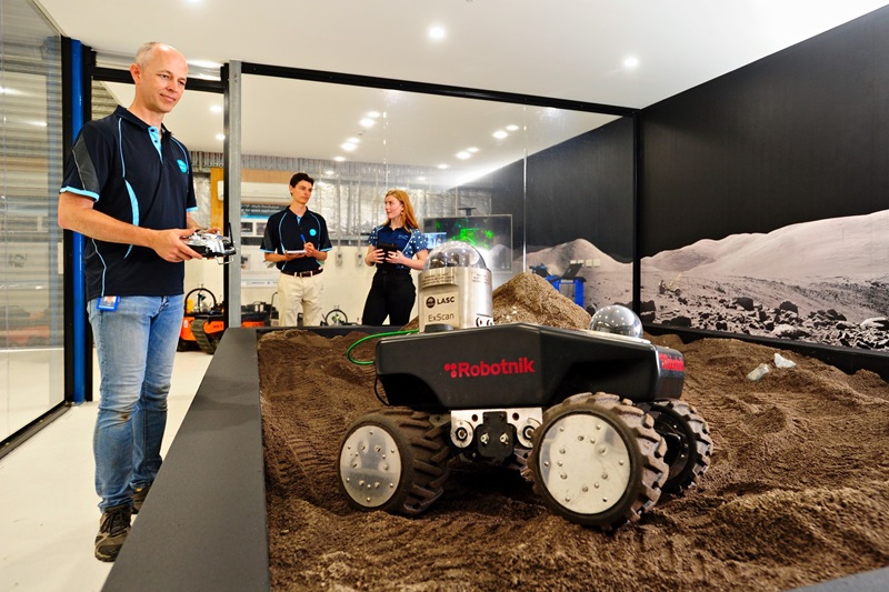 Man operating remote control vehicle over simulated moon dust, watched by man and woman