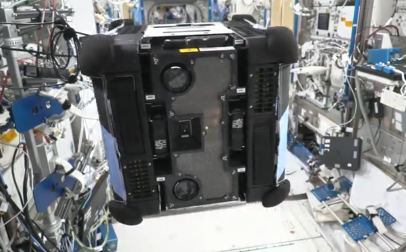 a floating cube in the foreground with two lenses. the background show lots of wiring, tubing, equipment and technology attached to all the walls. 