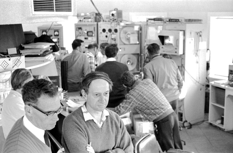 A black and white photograph of two men in the foreground looking at something to the side of the camera; in the background six men with their backs to the camera are looking at a bank of controls and switches.