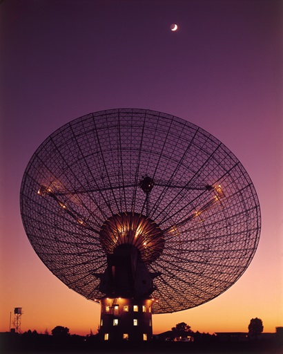 A large radio telescope dish, with the Moon above, at sunset; the sky is golden yellow at the horizon and fades to a deep purple at the top of the photo.