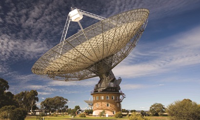 A large radio telescope antenna with green grass and small shrubs in the foreground and blue sky and white clouds in the background.