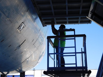 Image shows a man in a yellow hi-vis vest standing on a raised platform. The man is working on a aircraft fuselage. 