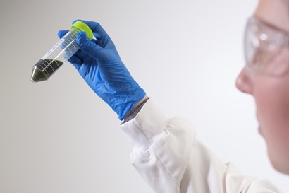 A female stands slightly out of frame, what we see of her face is out of focus. She is using her right hand to hold up a clear plastic vial that contains a dark green liquid. The vial is a cylinder with a tapered end, it has a bright green lid, and is about the same length as her hand. She is wearing safety glasses, a white lab coat, and blue latex gloves. 