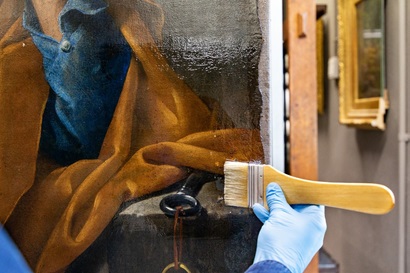 Image shows a gloved hand applying a clear resin to an artwork with a wooden and bristle brush. The sections of the artwork with resin appear wet. The artwork shows folds of fabric in brown and blue colours. In the background other images hang on the wall suggestive of an art gallery.