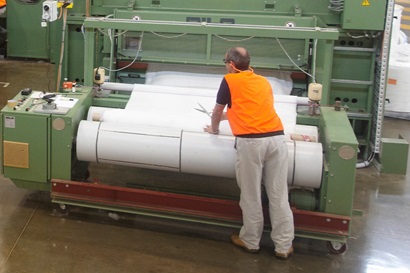 A man wearing an orange hi-visibility safety best stands at an machine that is rolling large bolts of white fabric.