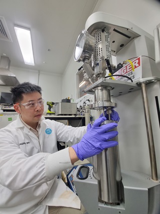 male scientist in white coat inside a laboratory holding a metal flask underneath a white machine.