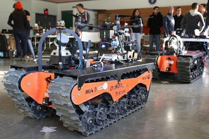 Two robots in a warehouse with people in the background preparing for the DARPA Subterranean Challenge. A drone rests on top of a large tank-like bot.