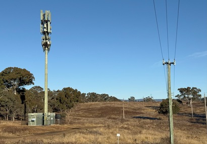 Mobile phone tower in a dry rural landscape with power lines and scattered trees under a clear blue sky.