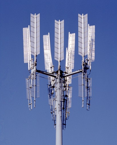 Close-up of a telecommunications tower with multiple rectangular panel antennas mounted in clusters at the top, set against a clear blue sky.