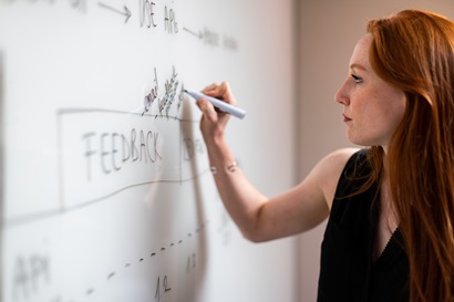 Woman with red hair and black shirt writes on a whiteboard