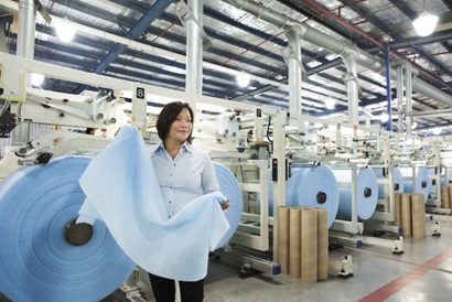 A woman stands in a textile factory. She appears in the left hand side of the foreground, looking out of shot to the right. She holds a length of light blue fabric in both hands. She is standing in front of a large roll of what appears to be the same fabric. The roll of fabric is the same height as her torso. In the background of the shot we can see many rolls of fabric on machinery.