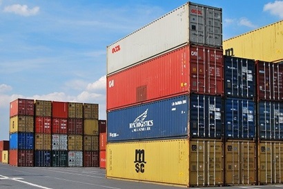 Brightly coloured shipping containers sit stacked on a dock. The containers in the foreground are stacked four high. The containers are blocks of yellow, blue, red and grey.