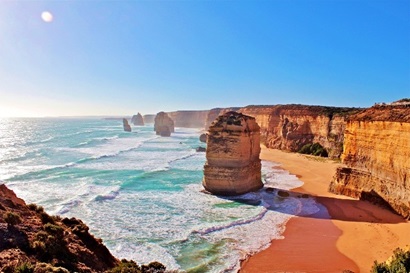 Image is a landscape view of the 12 Apostles. The sea and sky are glue, while the cliff face is a warm yellow orange.
