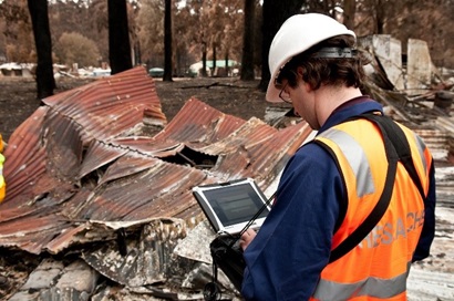 A man is turned away from the camera, he is wearing a blue shirt and orange hi-visibility vest. He is using an e-devise and is standing in from of a burnt building.