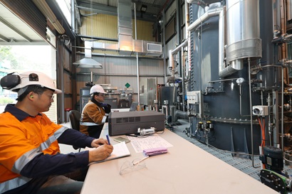 Two men in orange vests work at a table in a large factory
