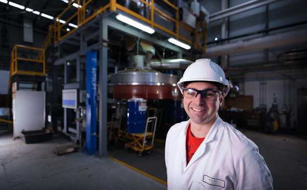 Male researcher, Jason Donnelly, wearing helmet, safety glasses and white lab coat, standing in front of Dry slag granulation rig.