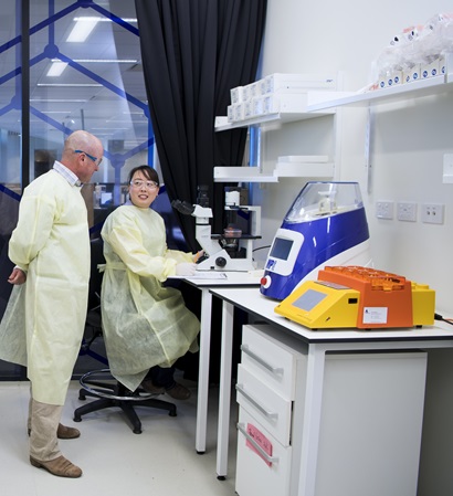 A male and female scientist wearing disposable lab coats work at a bench in a brightly lit lab with brightly coloured equipment around them.