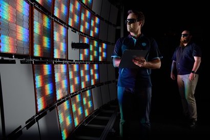 A person with a clipboard stands in front of a large curved wall of rectangular panels showing bright, rainbow-colored light pattern, with another person in the background. The setting is dark, emphasizing the vivid colors on the panels.
